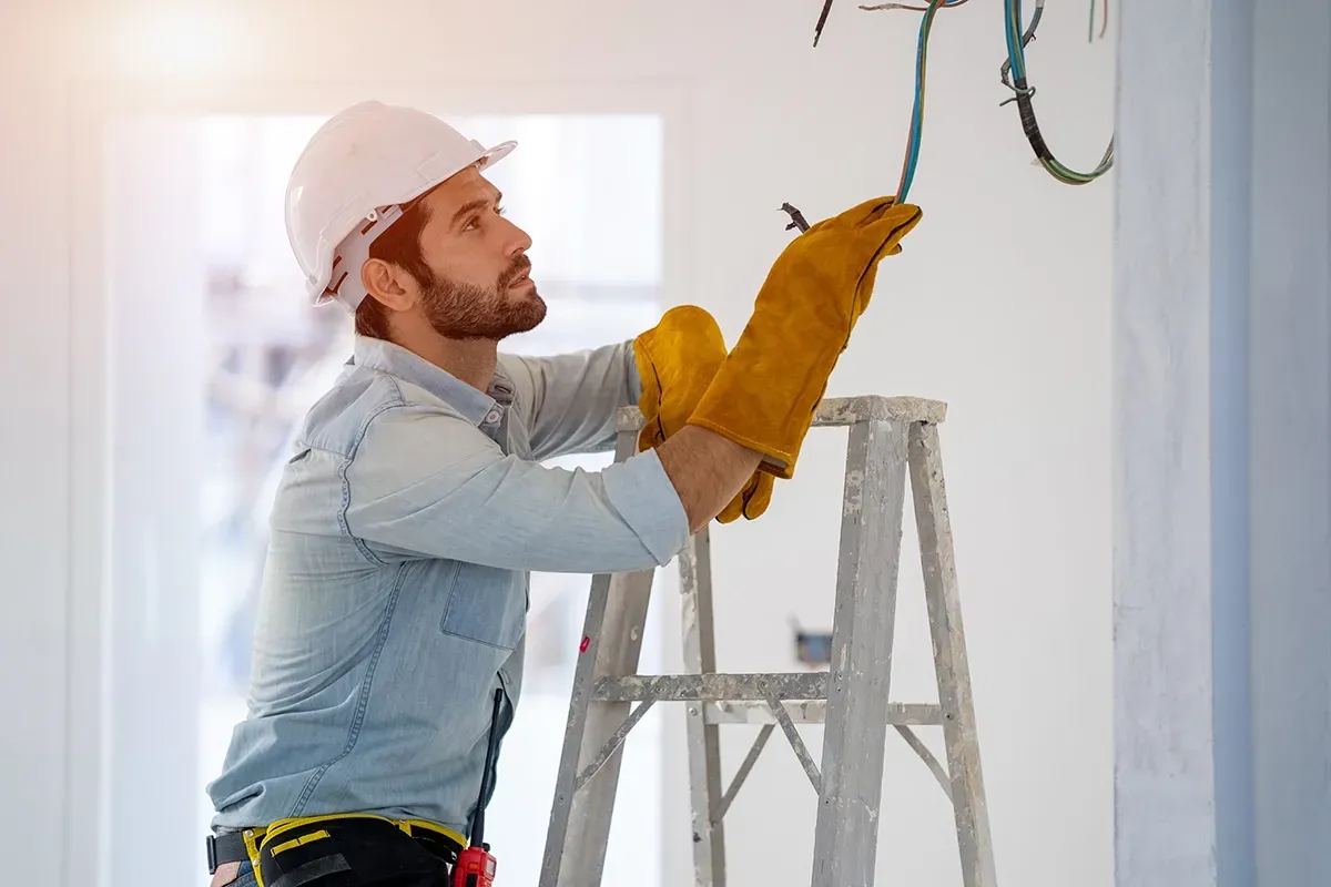 An electrician in a white hard hat on a ladder, gloved hands working on ceiling cables
